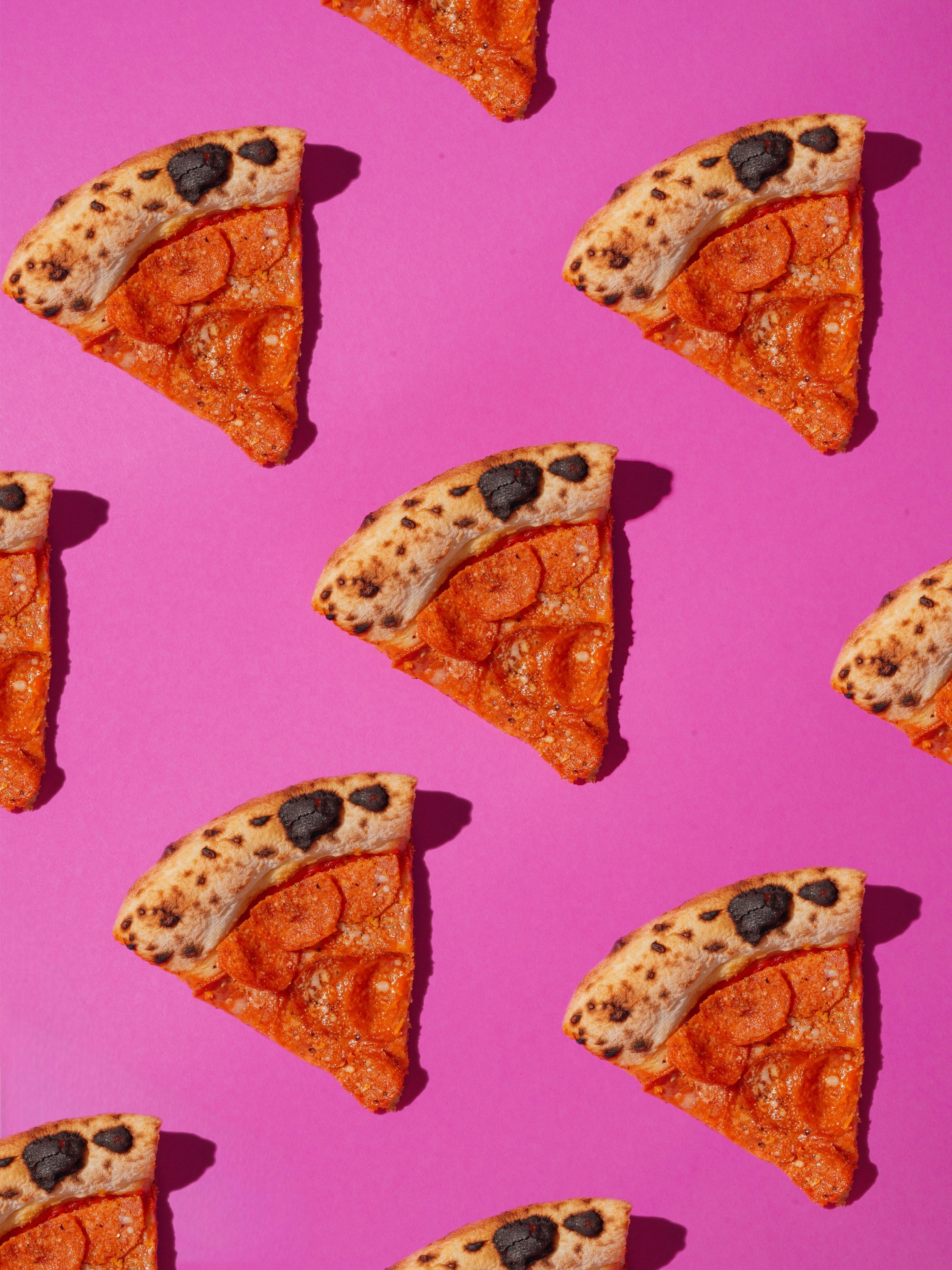 A pink background with a pattern of pepperoni and olive pizza slices - Pizza