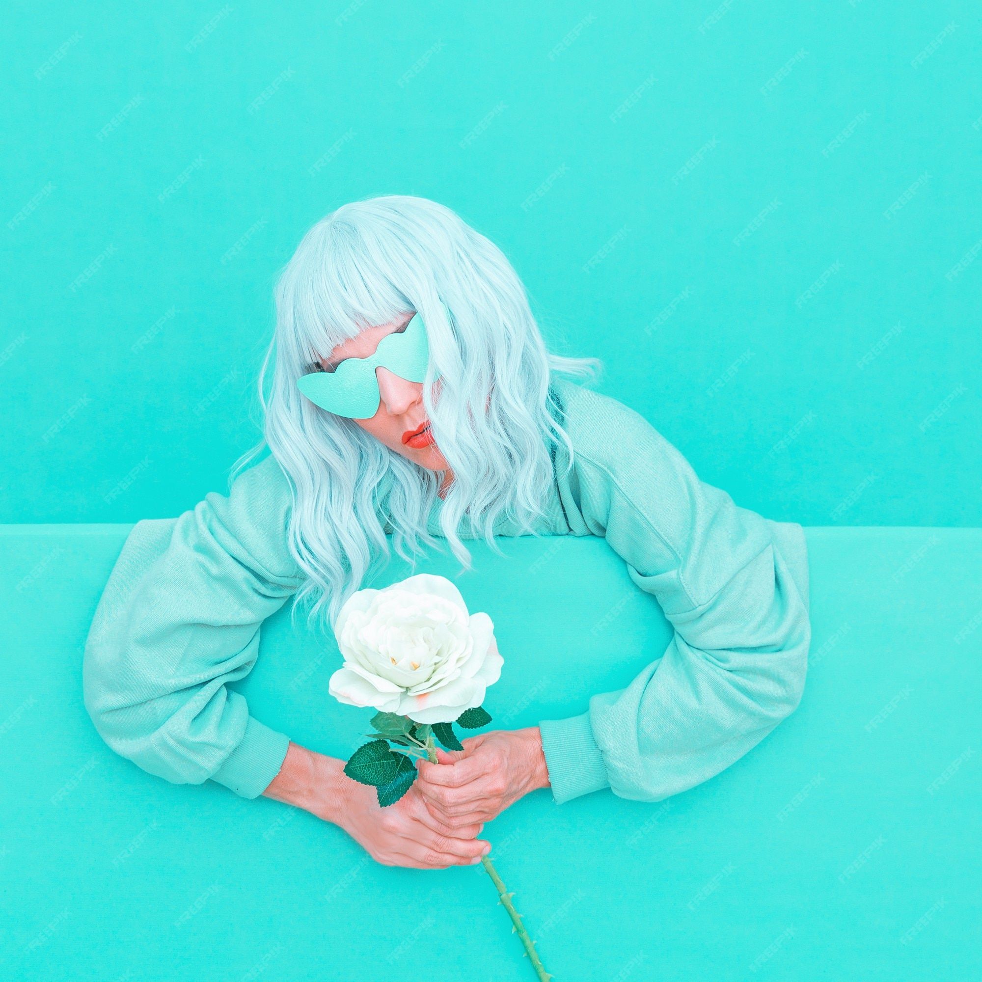 A woman with blue hair and sunglasses holding a white rose against a blue background. - Aqua
