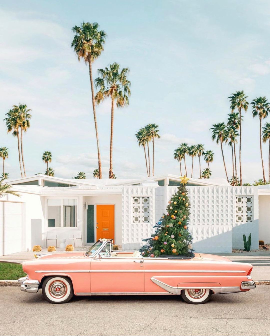 A pink vintage car with a Christmas tree on top parked in front of a white house with palm trees. - Retro