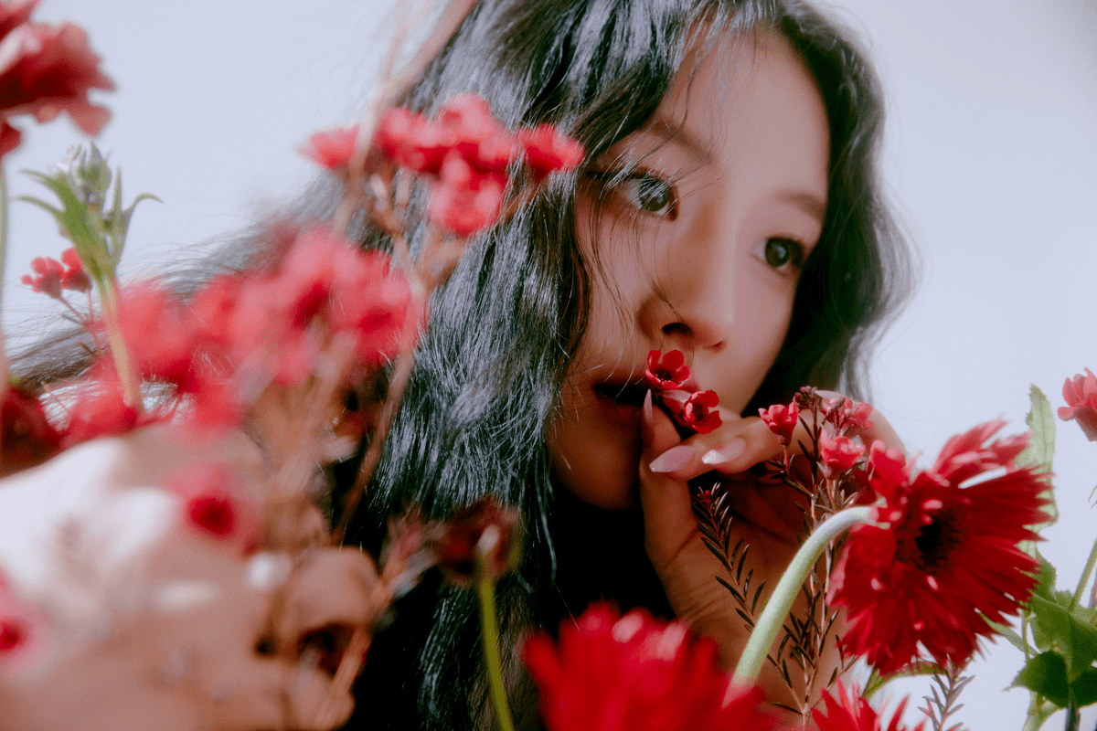 A woman with long black hair standing behind red flowers. - FIFTY FIFTY