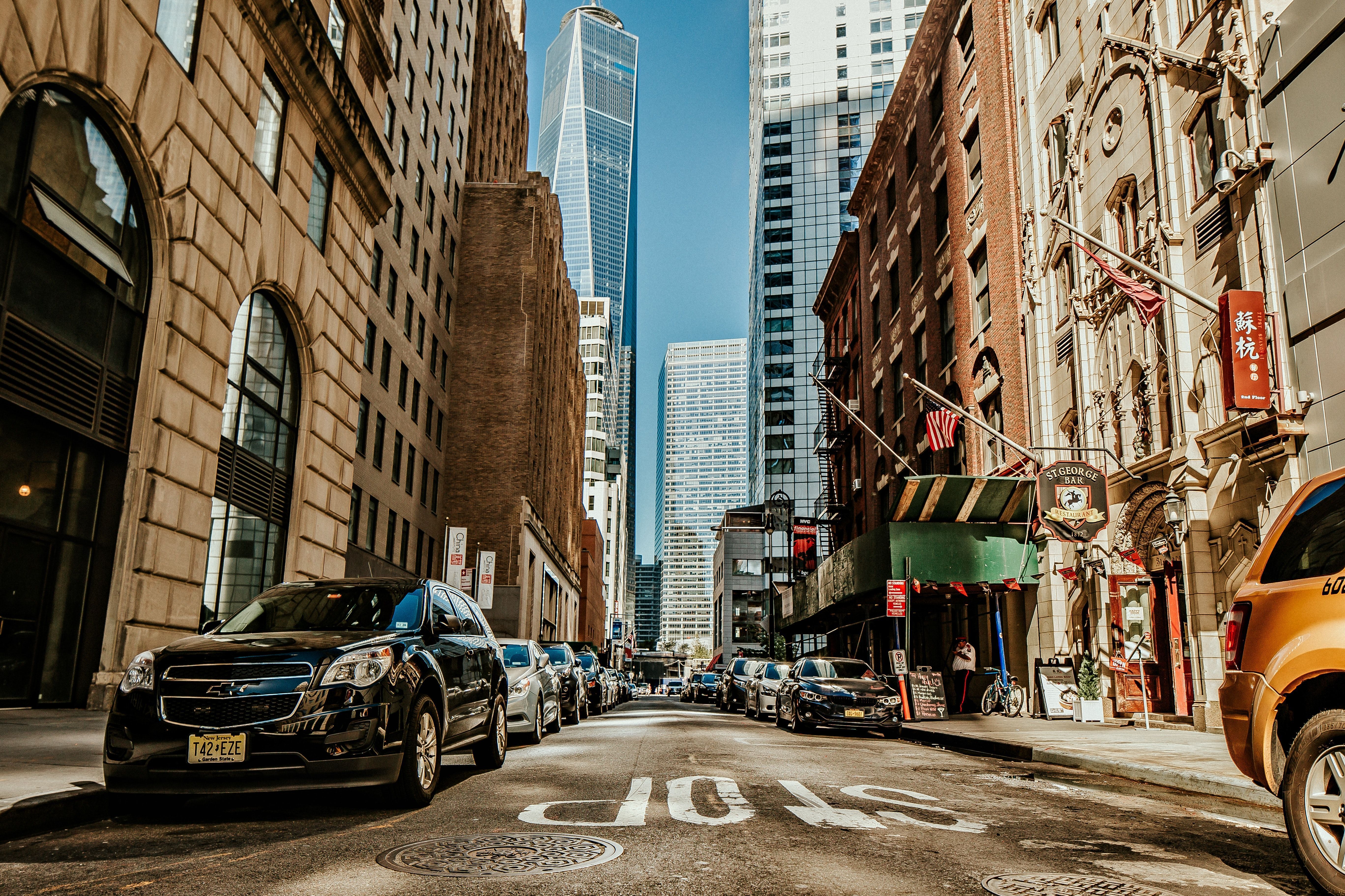A street in New York City with cars parked on the side and a skyscraper in the background. - New York