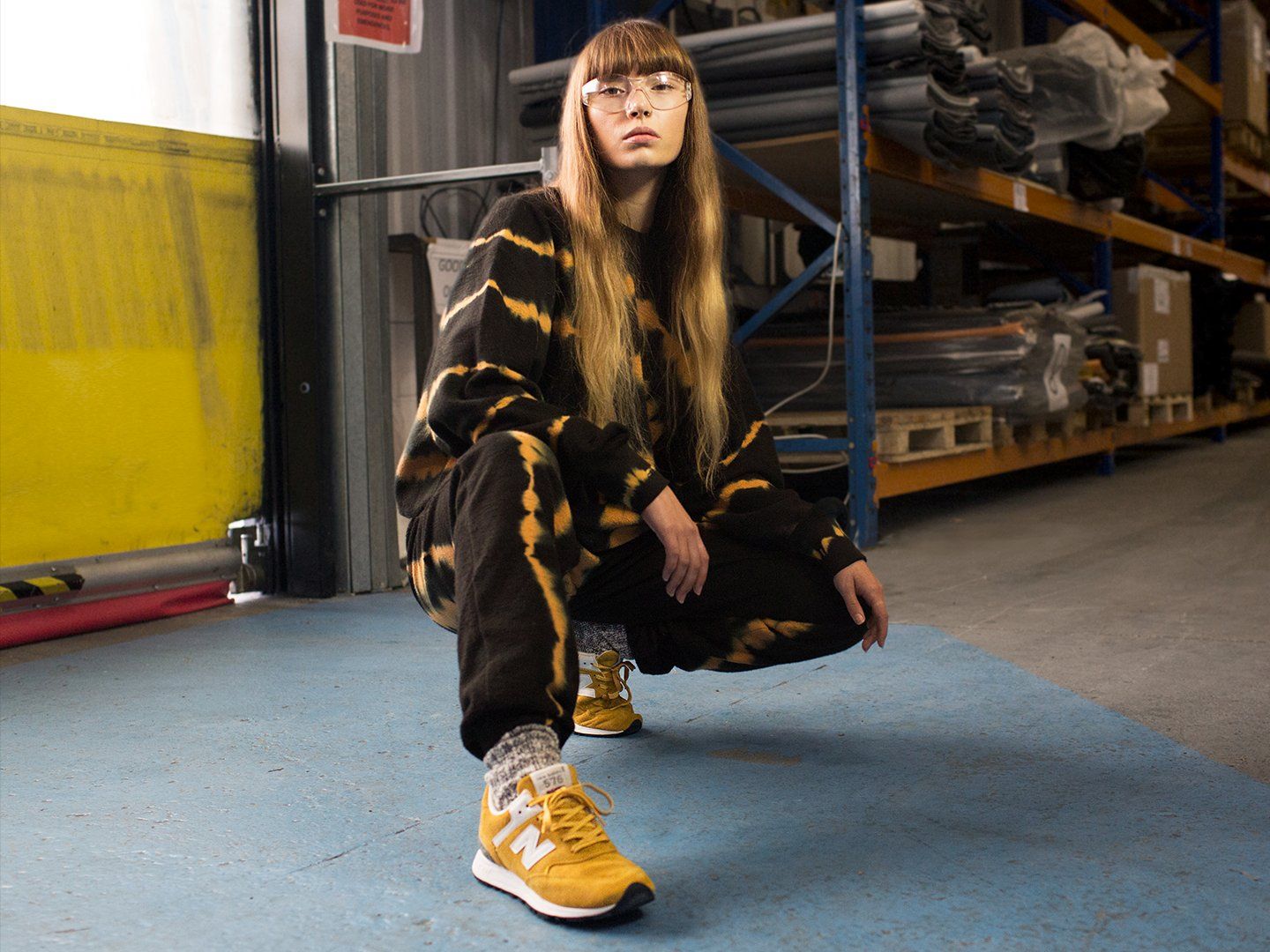 A woman crouching down in a warehouse wearing a black and yellow tie-dye sweat suit and yellow New Balance sneakers - New Balance