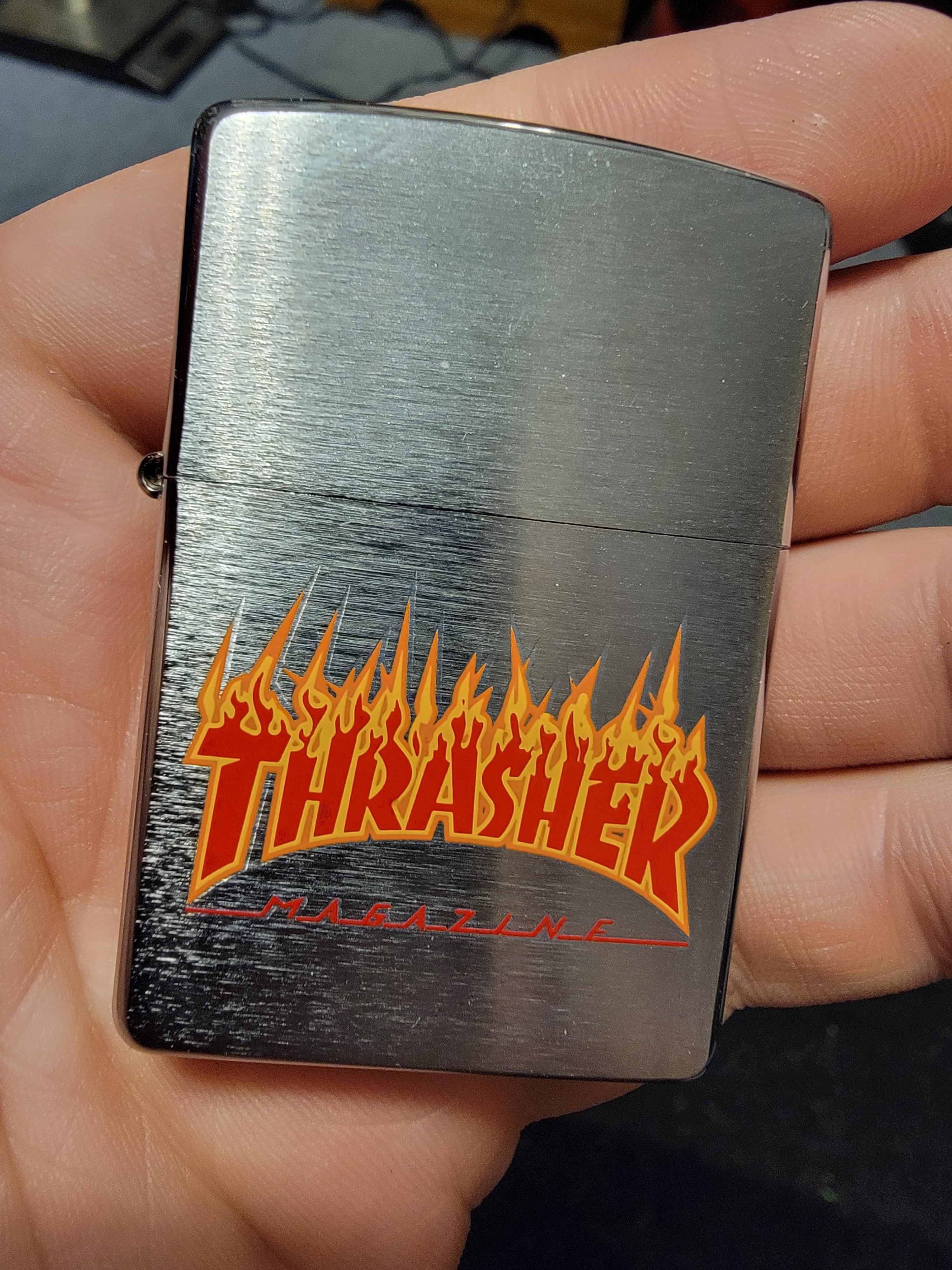 A hand holding a Zippo lighter with the Thrasher magazine logo on it. - Thrasher