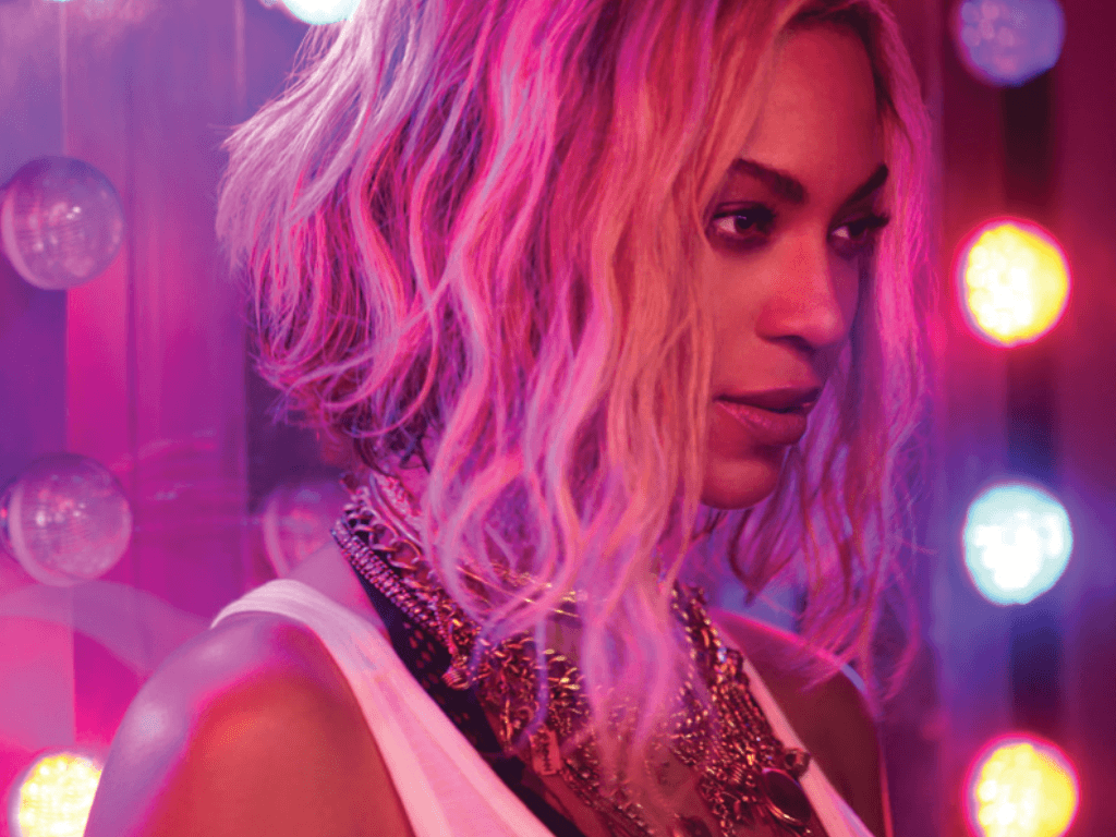 Beyoncé, wearing a white top and necklaces, poses in front of a wall of mirrors and pink and purple lights. - Beyonce