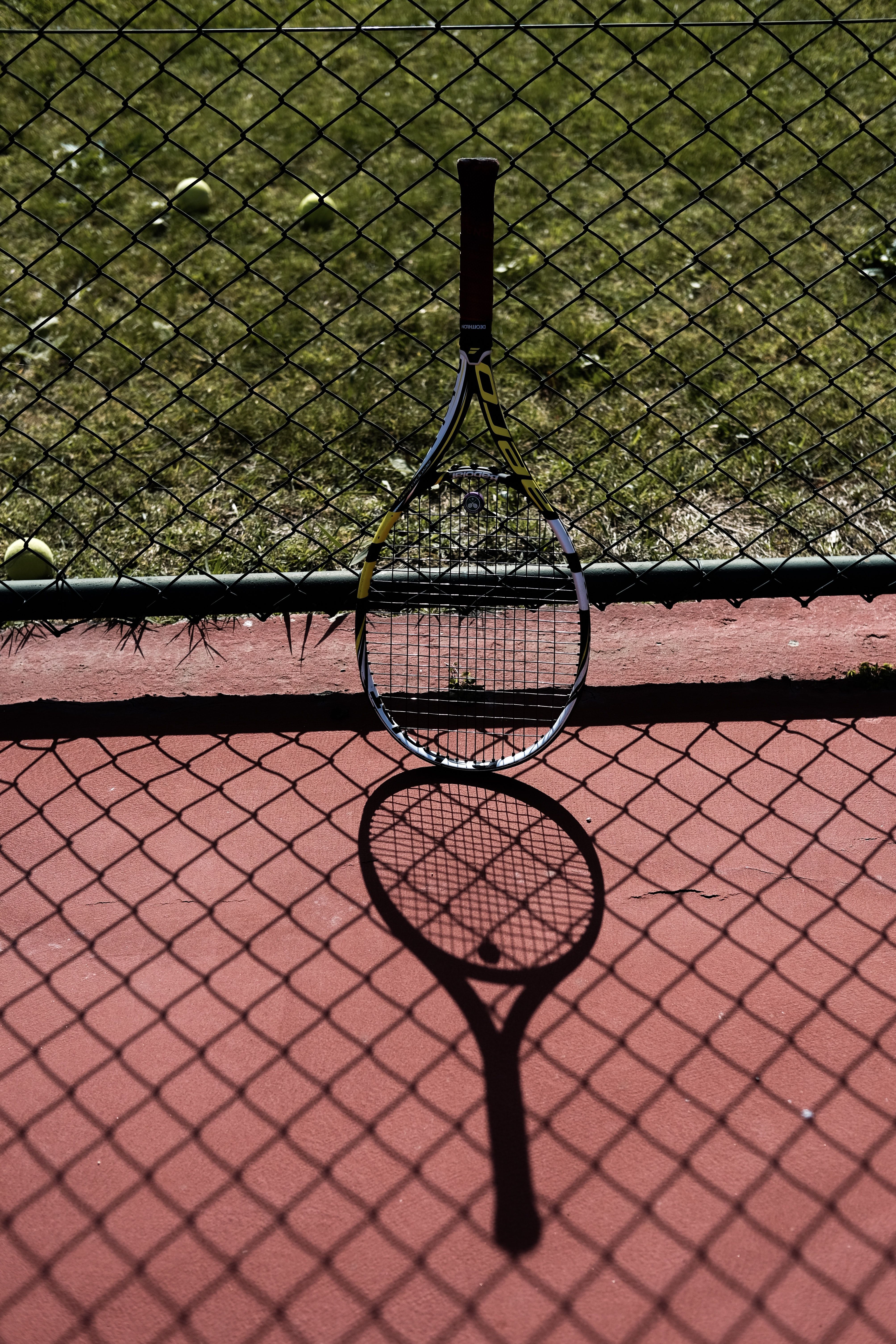 A tennis racket hanging on a fence with its shadow on the ground. - Tennis