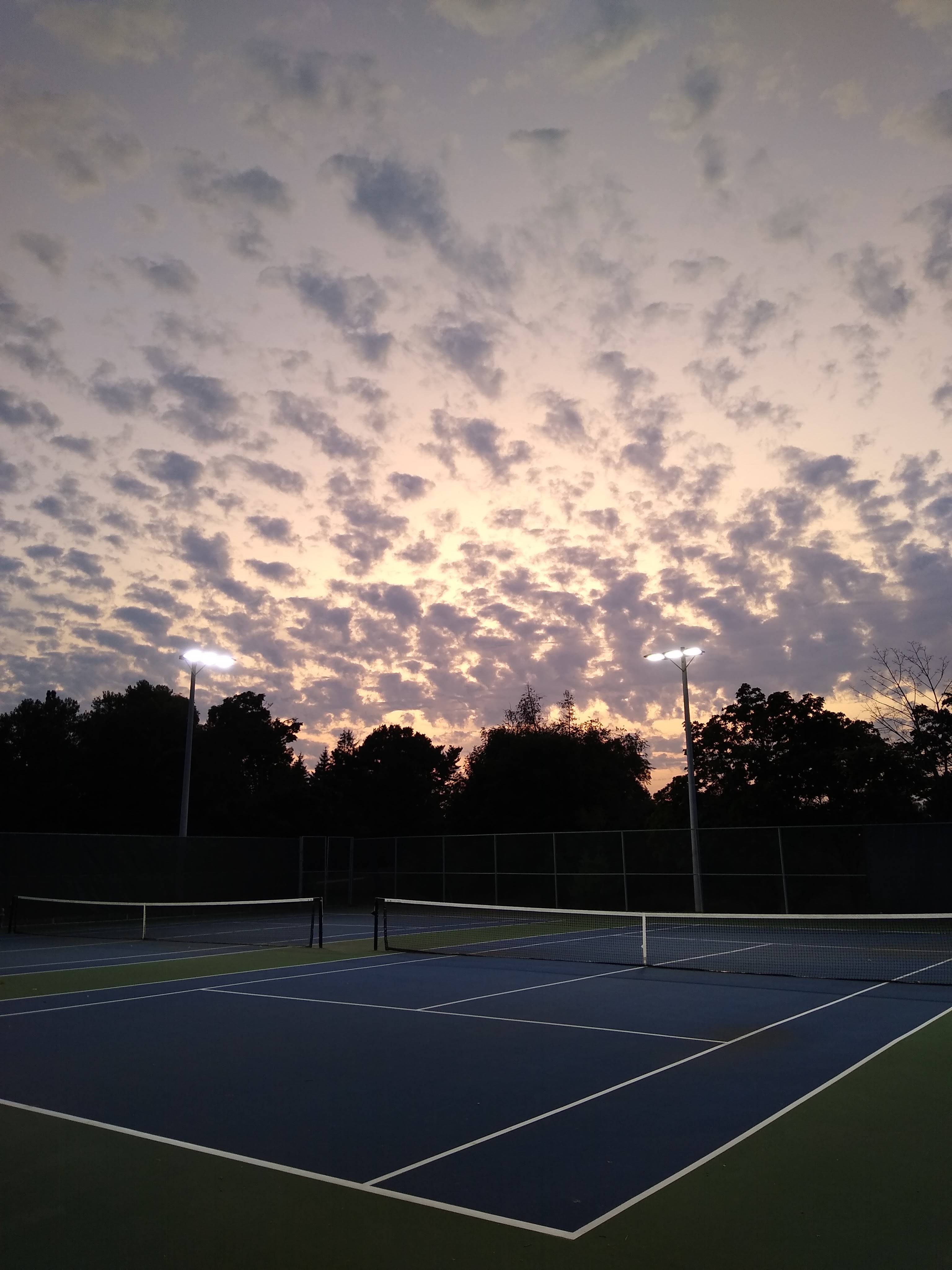 A tennis court with two lights on the side and a beautiful sunset in the background. - Tennis