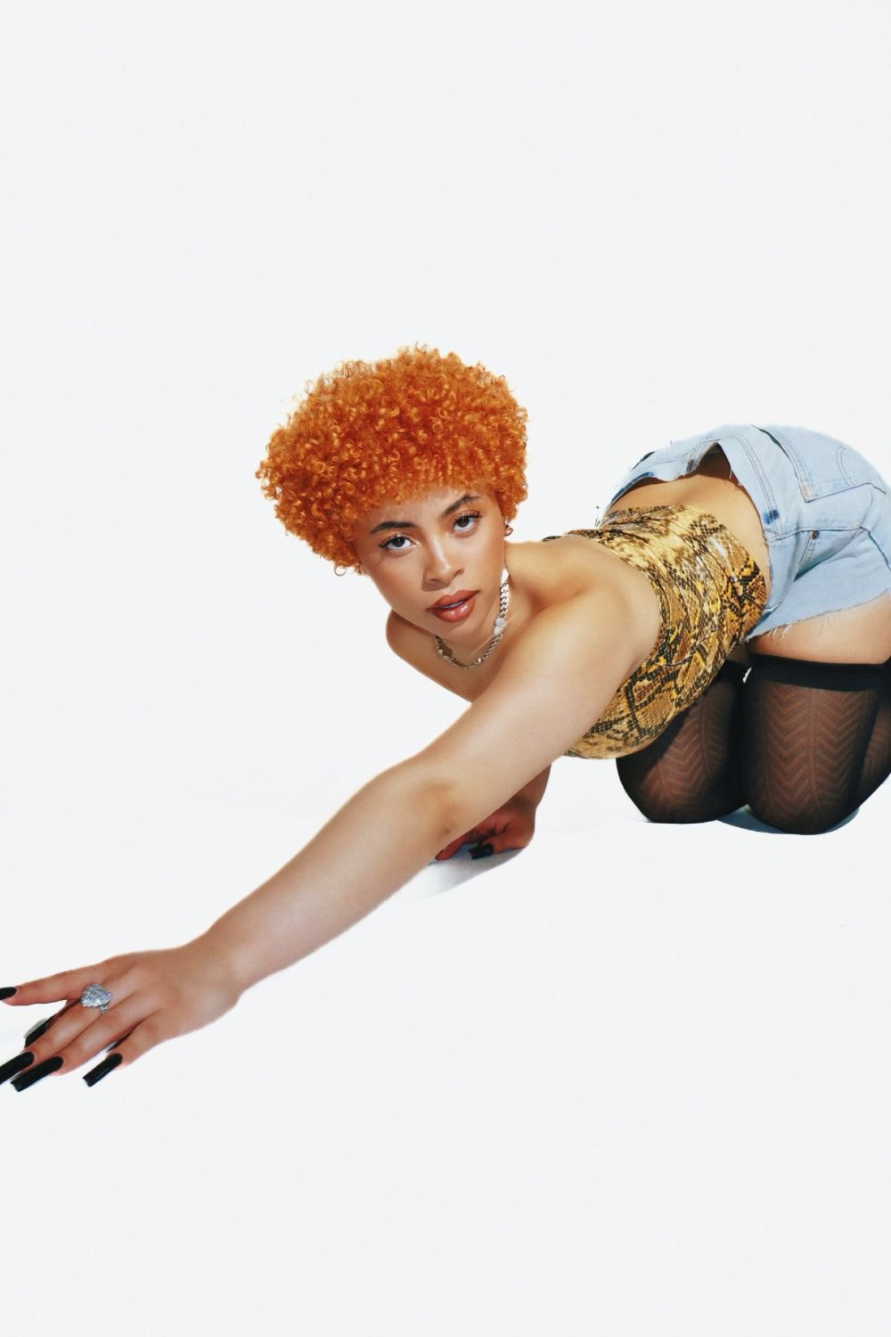 A woman with orange hair and a gold top leans over, touching the ground with her hand. - Ice Spice