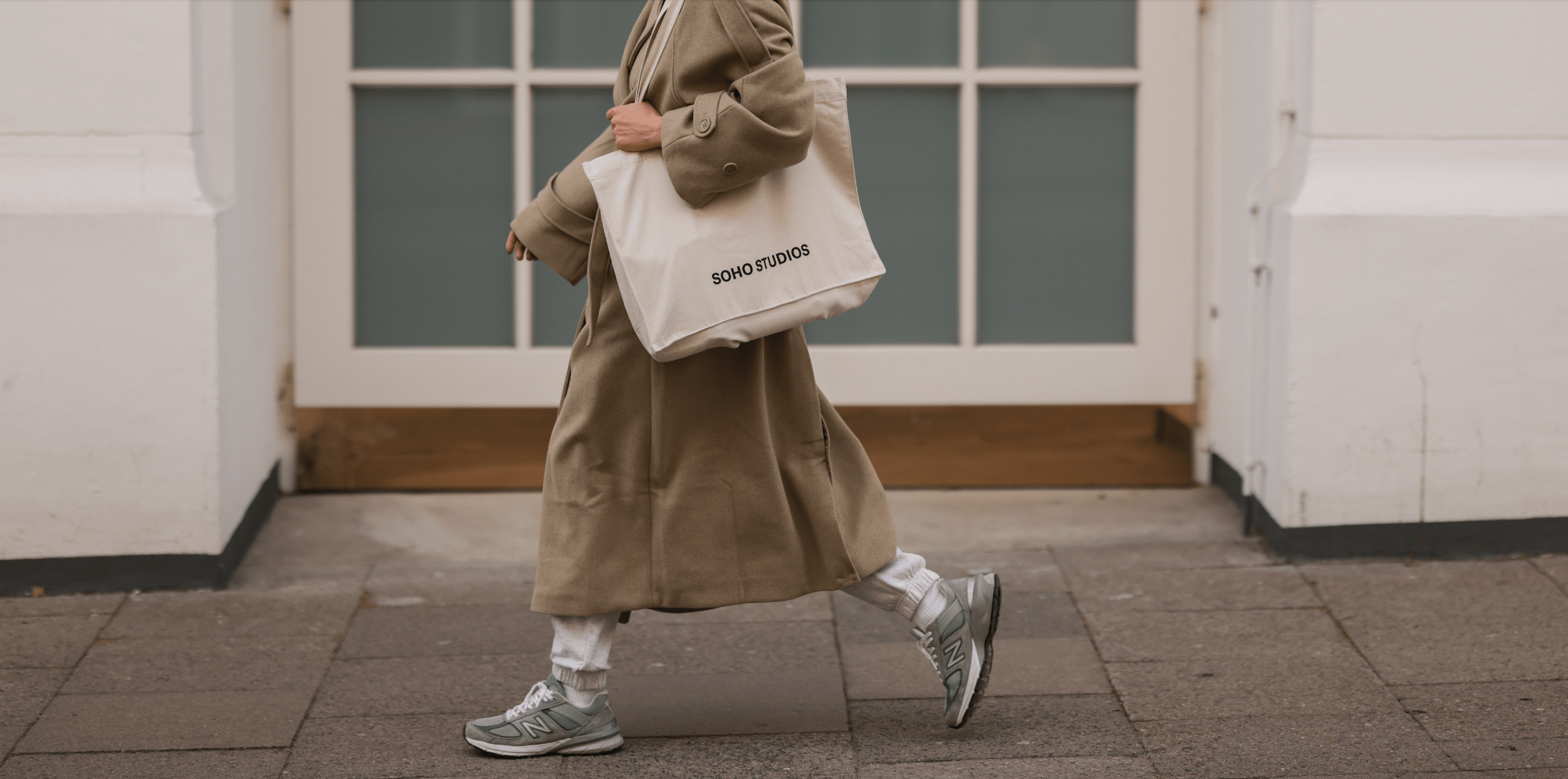 A woman wearing a beige coat and carrying a beige tote bag walks down a street. - New Balance