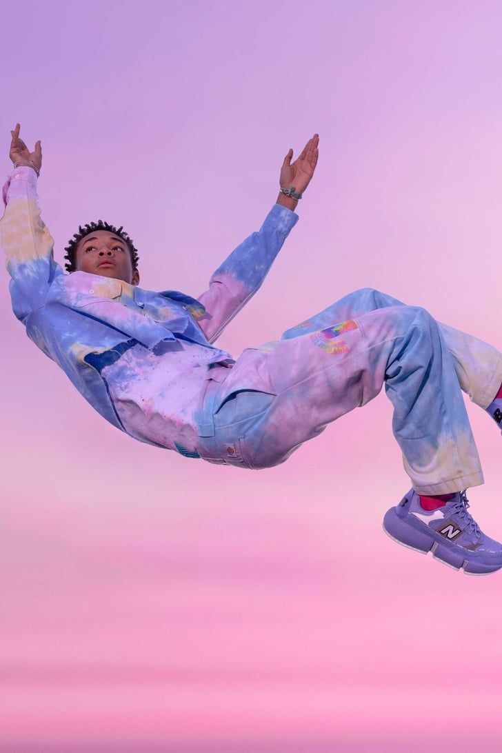 A model in a tie-dye sweatsuit and purple Nikes is mid-air, hands up, in front of a pink and purple gradient background - New Balance