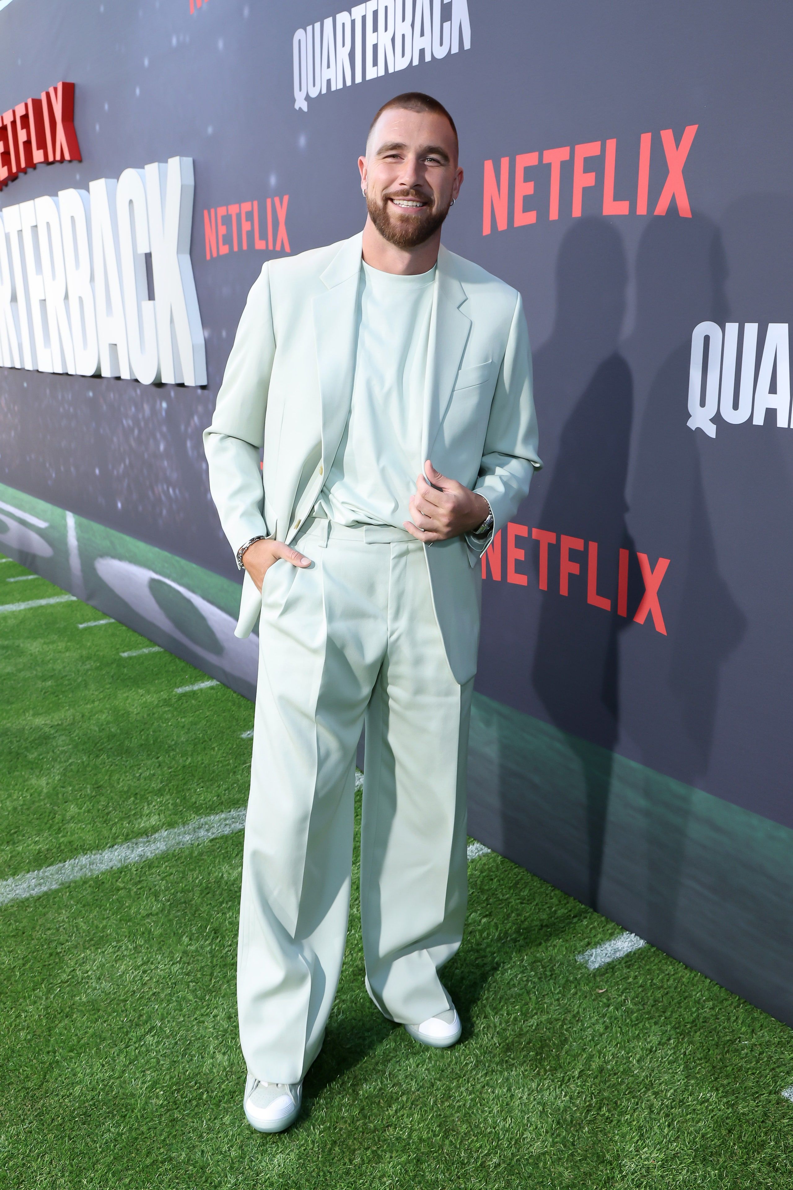 A man in a white suit standing on a green carpet - Travis Kelce