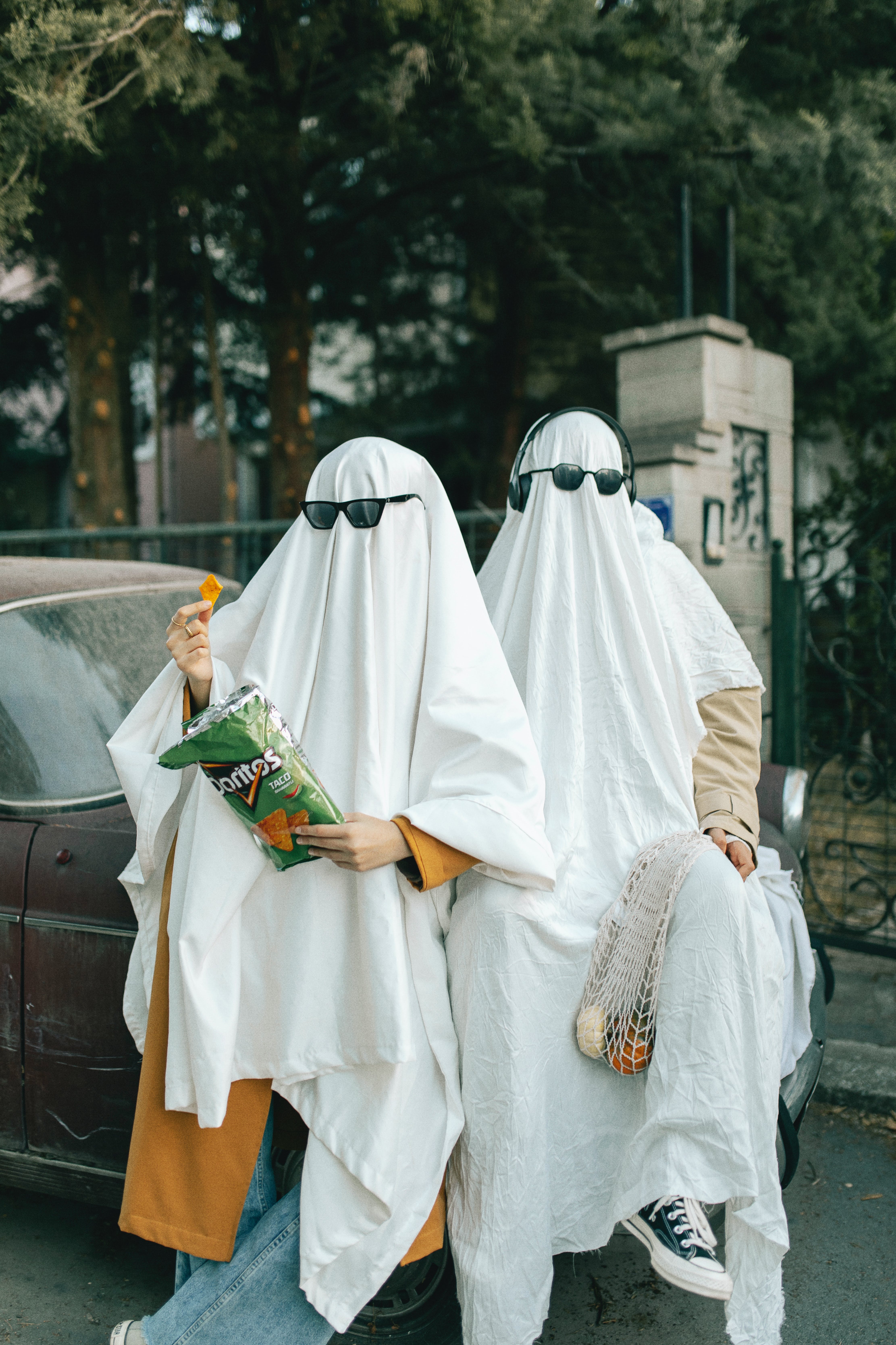 Two people in white sheets with sunglasses and snacks sitting on a car - Doritos