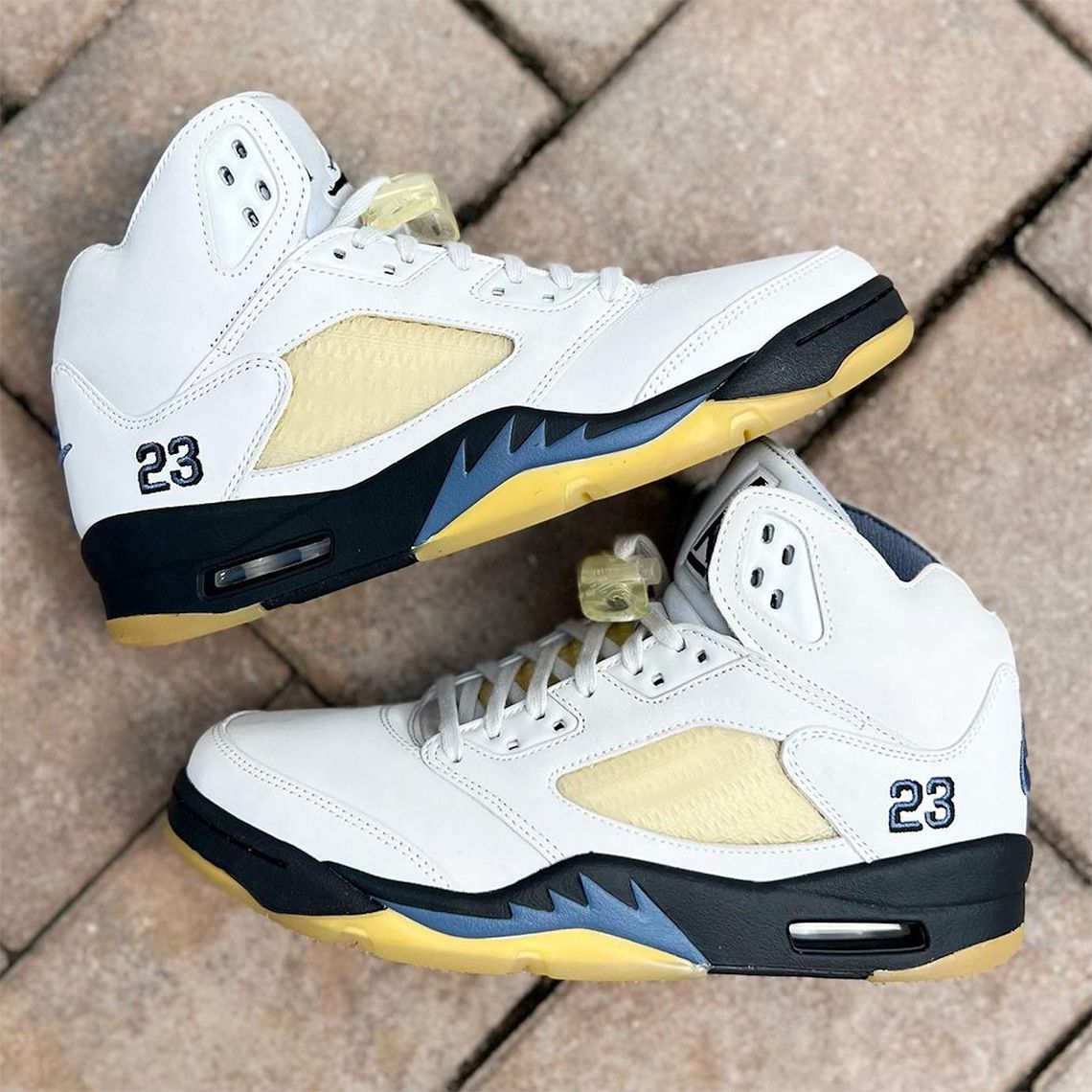 A pair of white Air Jordan 5 sneakers with yellow accents are shown on a brick surface. - Air Jordan 5