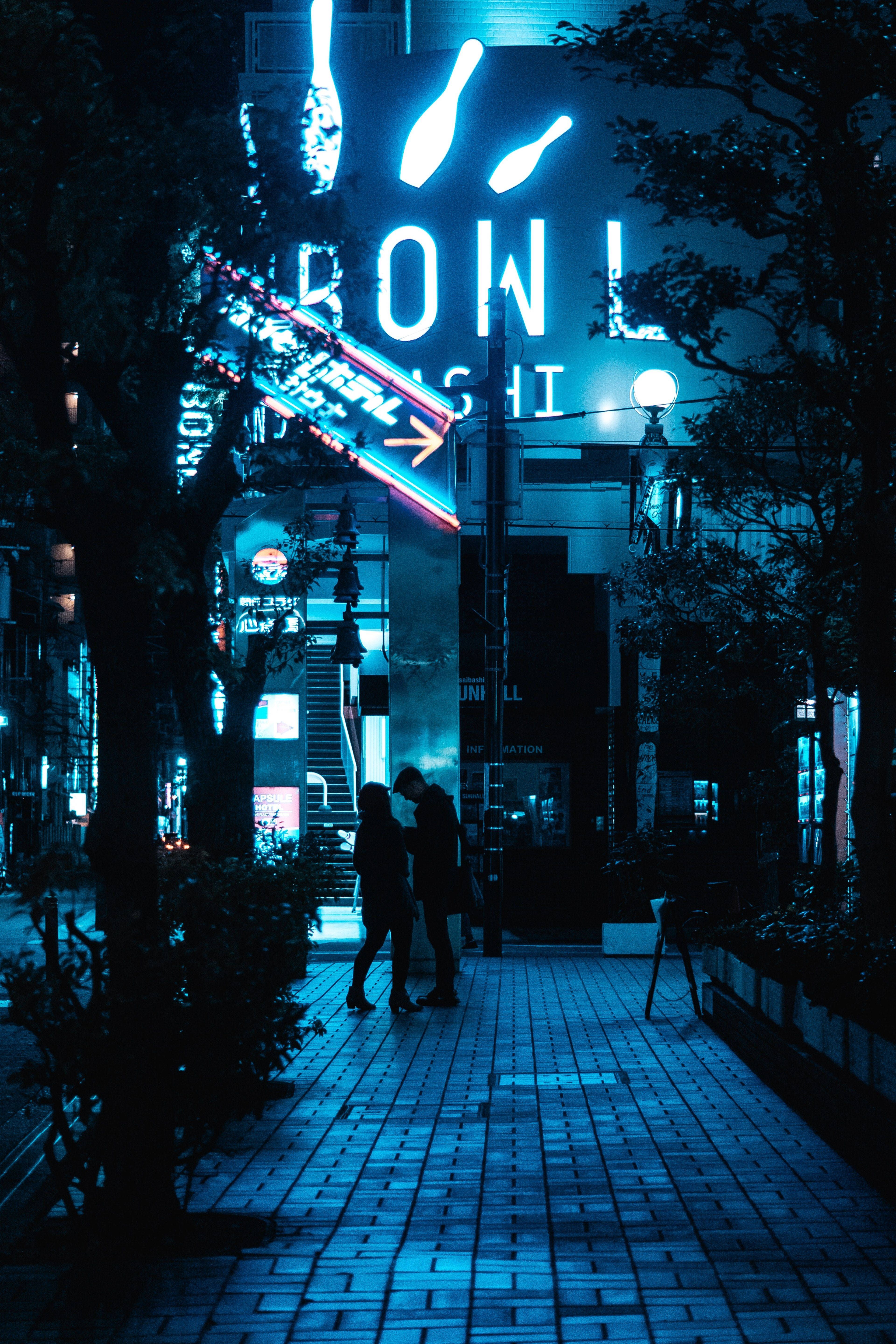 A couple walking down a street at night with neon signs in the background. - Navy blue, neon blue, dark blue, city