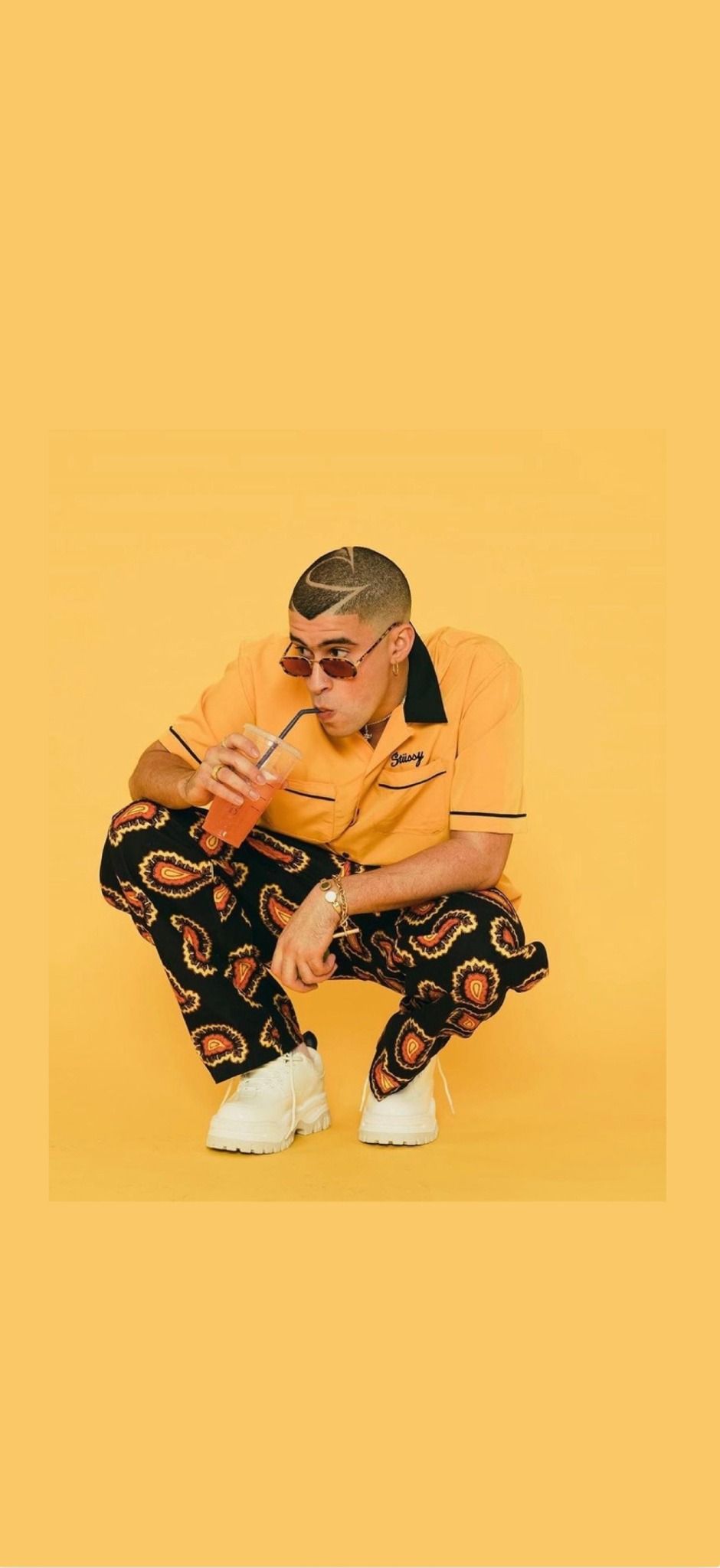 A man in a yellow shirt and black pants crouching down and drinking from a cup. - Bad Bunny