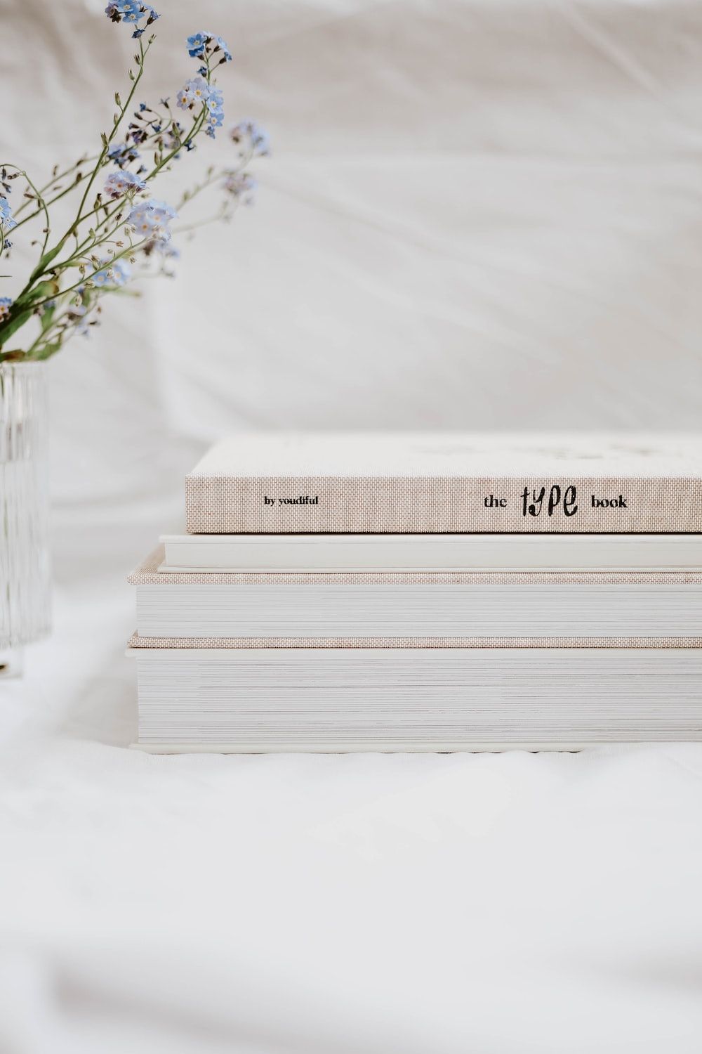 A stack of white books with a vase of flowers on a white sheet. - Books