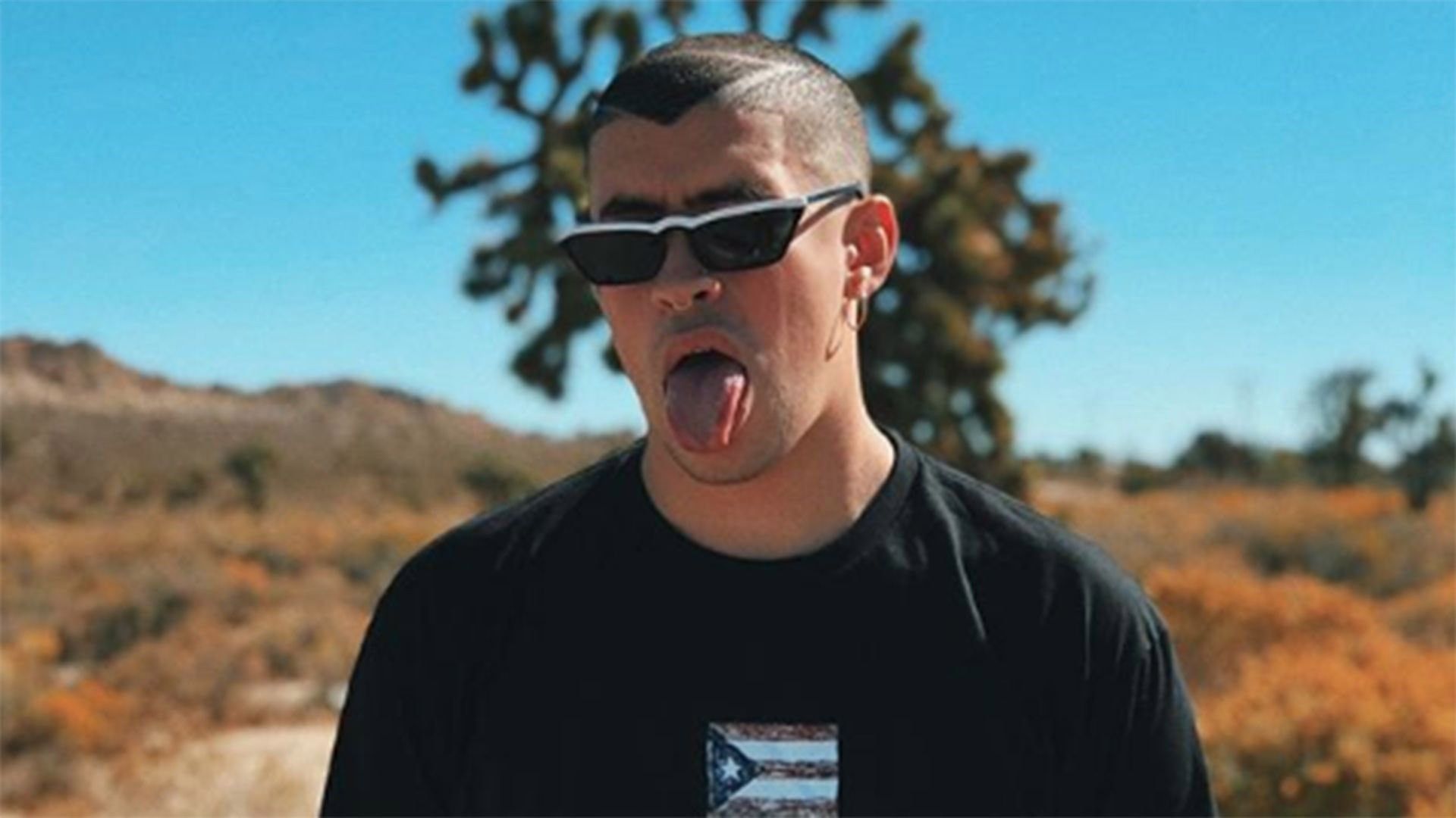 Aaron Marcellus, a white man with a shaved head, wears sunglasses and sticks his tongue out. He is standing in front of a desert landscape with Joshua trees in the background. - Bad Bunny