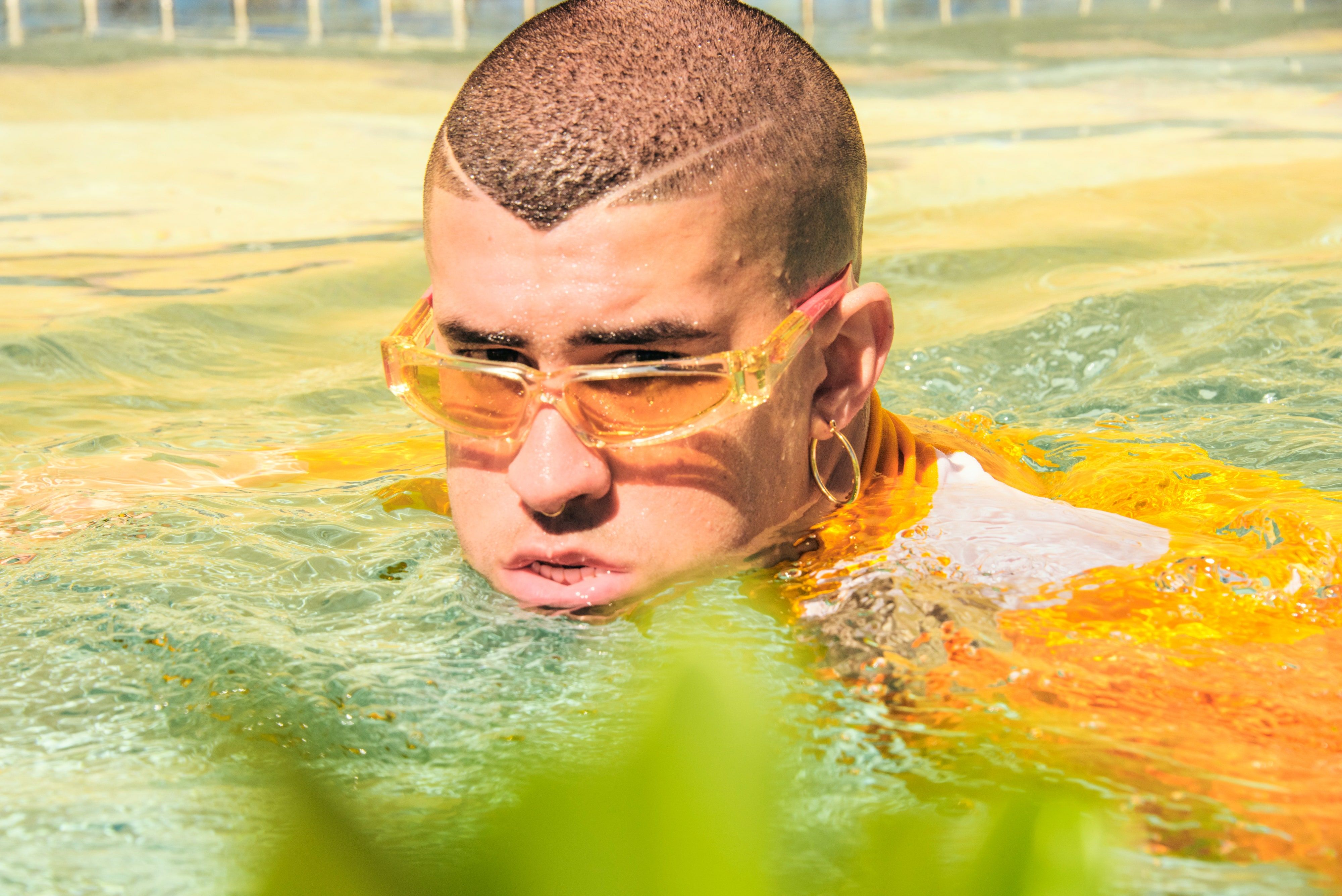 A man with a shaved head and wearing yellow sunglasses looks out from the water. - Bad Bunny