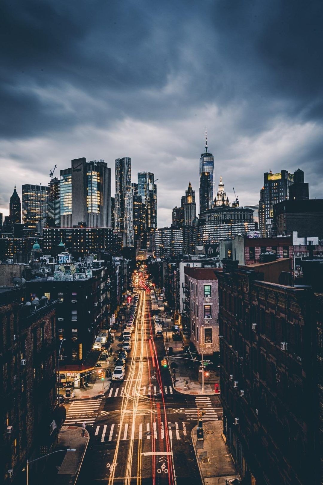 This image is of a cityscape at night with a very long exposure on the photo giving it a unique look. The street is filled with cars and the tall buildings have lights on in the windows. The sky is filled with dark clouds. - City, cityscape, New York, landscape, skyline