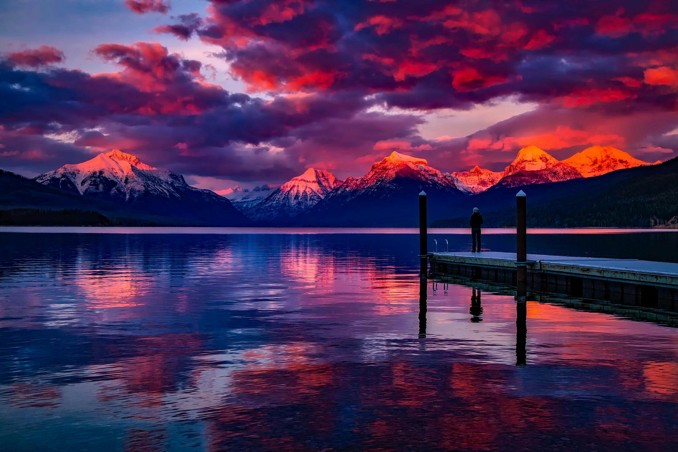 A man stands on a dock in front of a lake and mountains at sunset. - Desktop